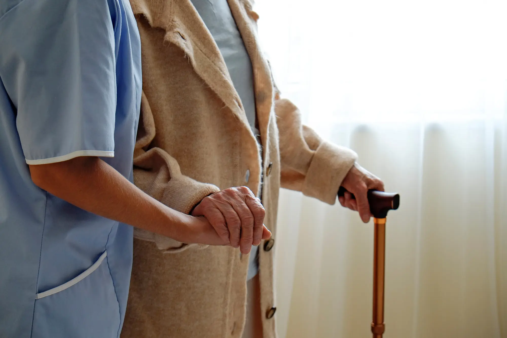Eine Pflegekraft hält liebevoll die Hand einer älteren Person mit Gehstock am Fenster